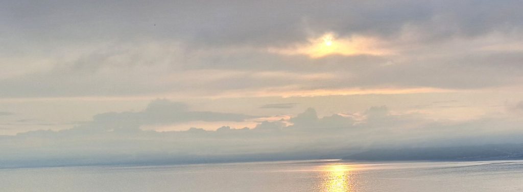 Ein bewölkter Himmel über dem Meer an der Küste bei Seatown, ganz in der Nähe des Meditations- und Retreat Zentrum Bonhays in Dorset. Die Sonne bricht durch die Wolken und das spiegelnde Sonnenlicht bildet einen rotorangenen Streifen auf dem glitzernden Wasser.