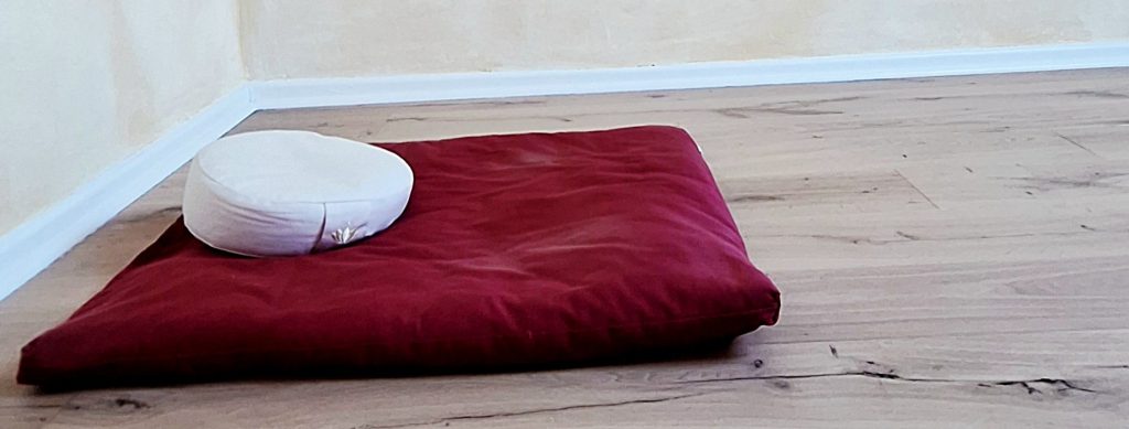 Ein weisses Zafu-Meditationskissen auf einem weinroten Zabuto-Mediationsunterlage. In einem Raum mit Fußboden aus Eichendielen und schlichten cremefarbenen Wänden. Das Bild symoblisiert Leere, Stille, innere Ruhe und Gelassenheit.